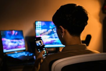 Person holding a smartphone in a dark room lit by glowing computer screens