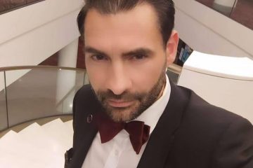 Томас Миллер в смокинге и бордовой бабочке на фоне лестницы в холле отеля