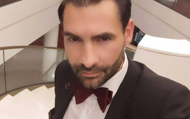 Томас Миллер в смокинге и бордовой бабочке на фоне лестницы в холле отеля