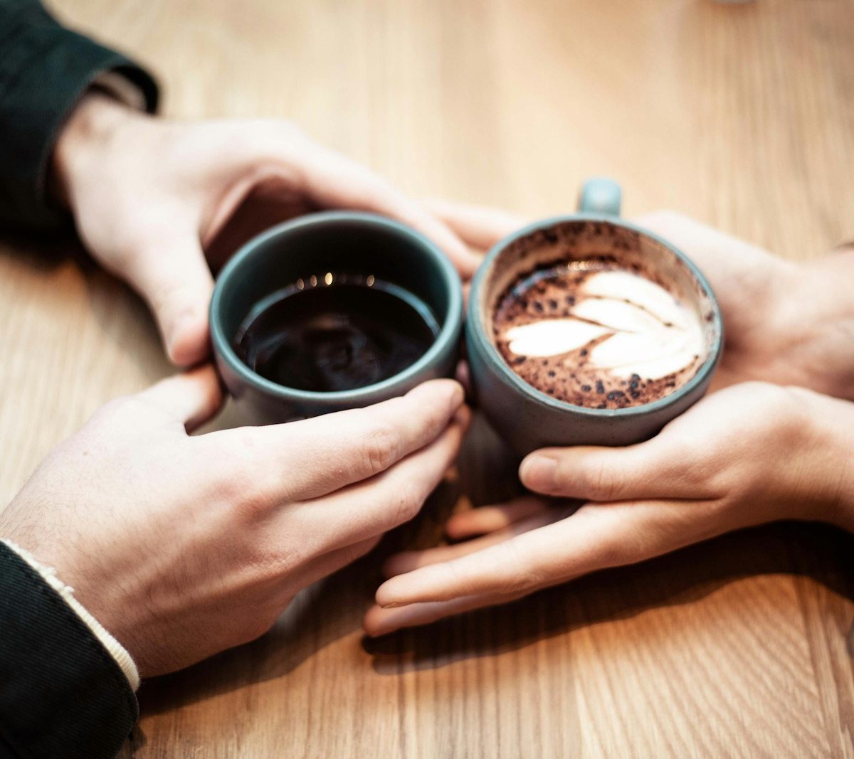 Two people's hands holding ceramic mugs of coffee across a table