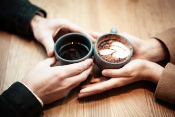 Two people's hands holding ceramic mugs of coffee across a table